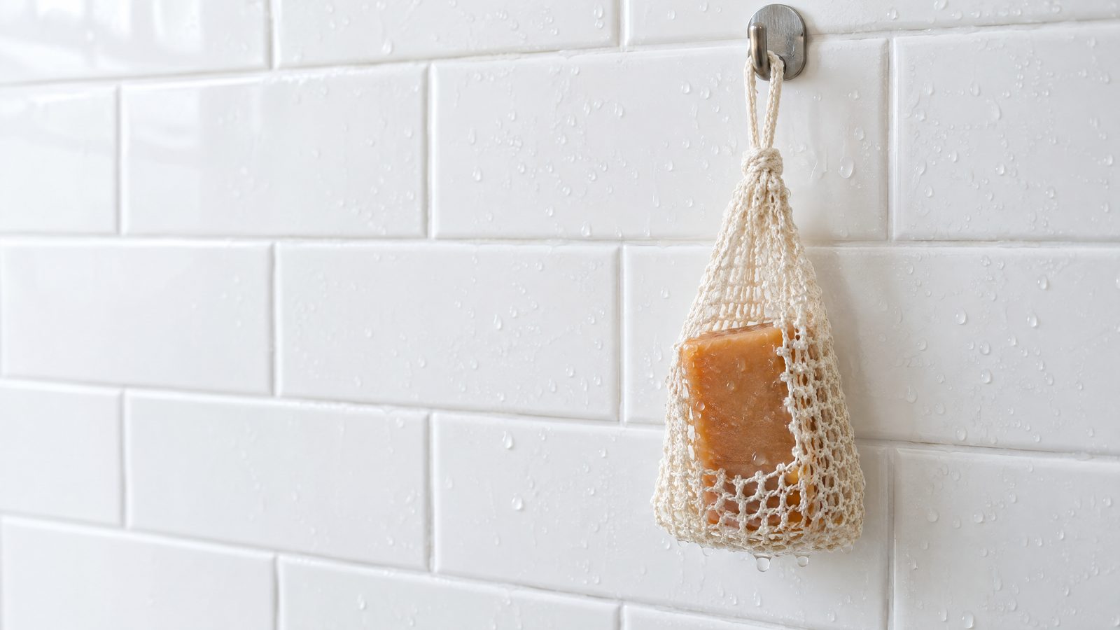 A persimmon soap bar in a mesh soap-saver bag hanging from a hook on white shower tile