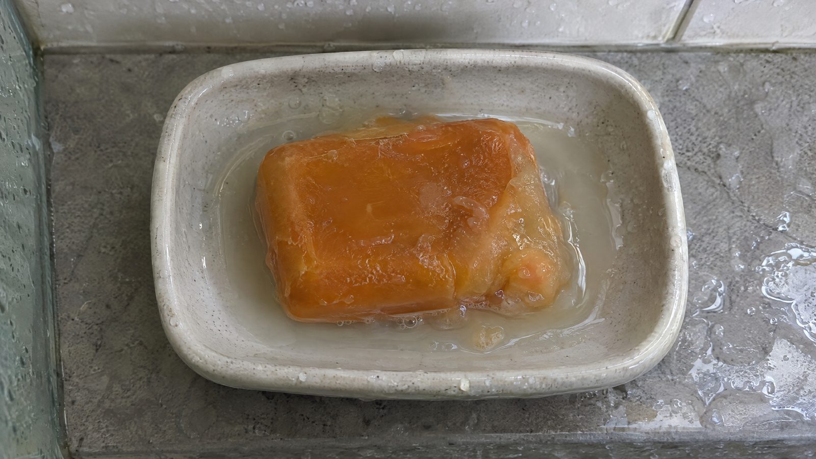 A softened orange-brown soap bar melting in a puddle in a ceramic soap dish